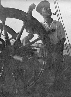 A photograph of Atch, tobacco pipe in mouth, grasping a ship’s steering wheel.