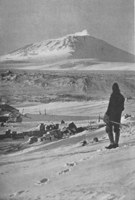 A photograph of a man holding a pickaxe with his back to the camera, facing Mt. Erebus in the distance.