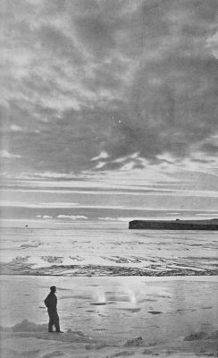 A photograph of a man standing on snow, facing a rocky outcropping in the distance.