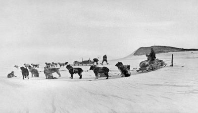 A photograph of a man on a sled with many dogs hitched to it. Another sled is ready in the background.