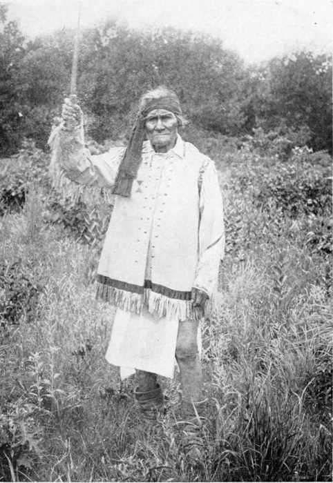 A photograph of an older Geronimo dressed in a fringed tunic and headband, holding a sword up in his right hand.
