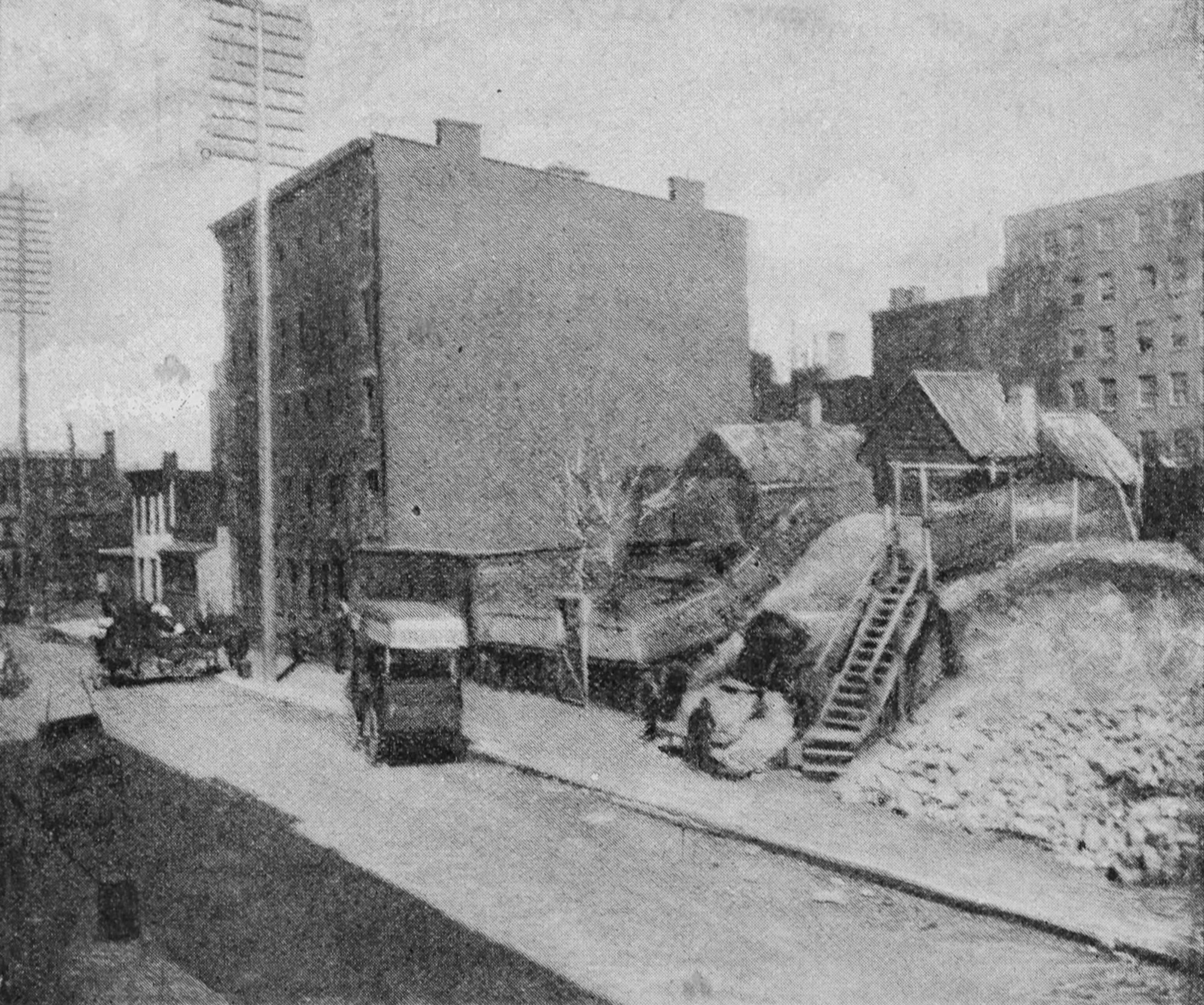 A street is shown with a couple of vehicles and some tall telegraph poles. A five-story building on the left has windows along the front but none on the side. Another multistory building is in the background on the right. There are few small dwellings nearby.