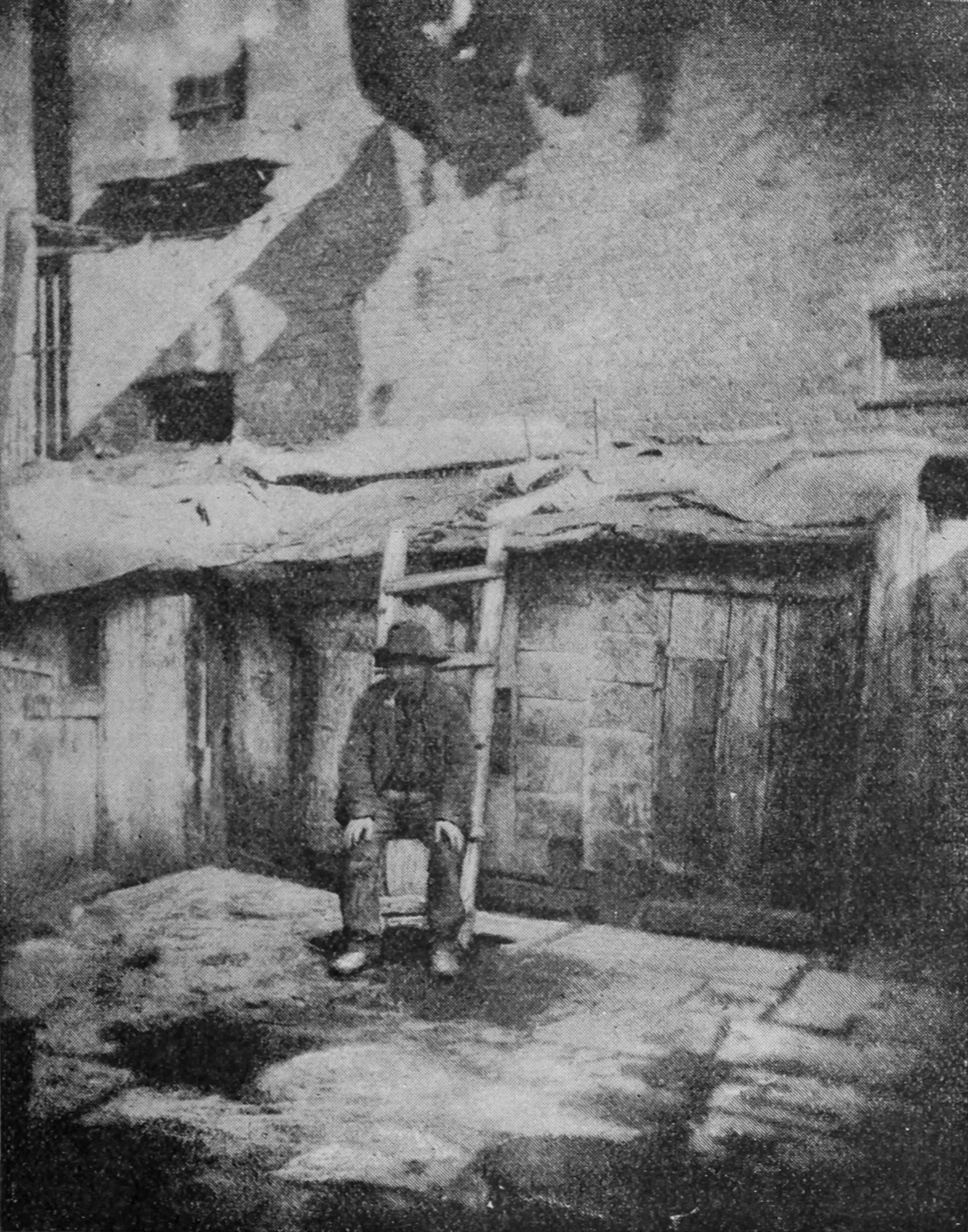 A man with a pipe in his mouth sits on a short ladder leaning against a shed at the back of a building.
