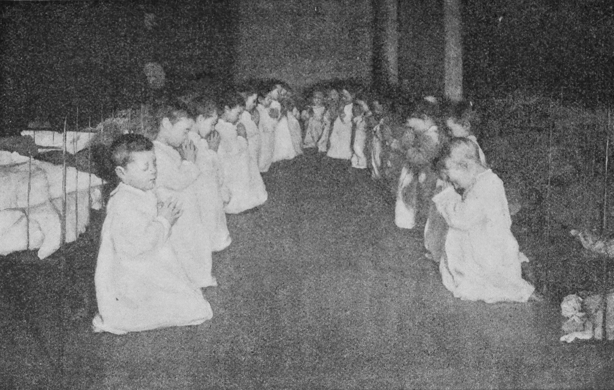 Children, kneeling in a semicircle, have their hands folded in prayer.