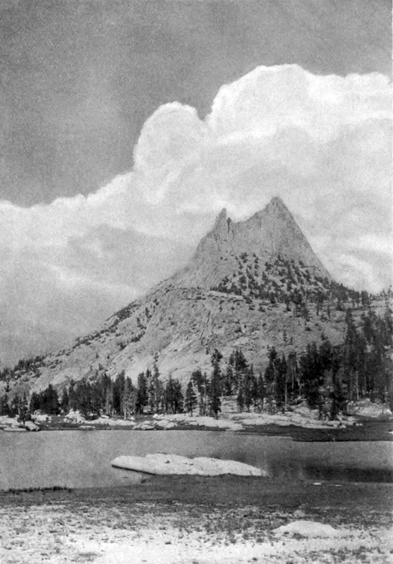 A distant, isolated mountain peak at the base of which lies a band of fir trees, a lake, and a small open beach covered in a thin layer of snow.