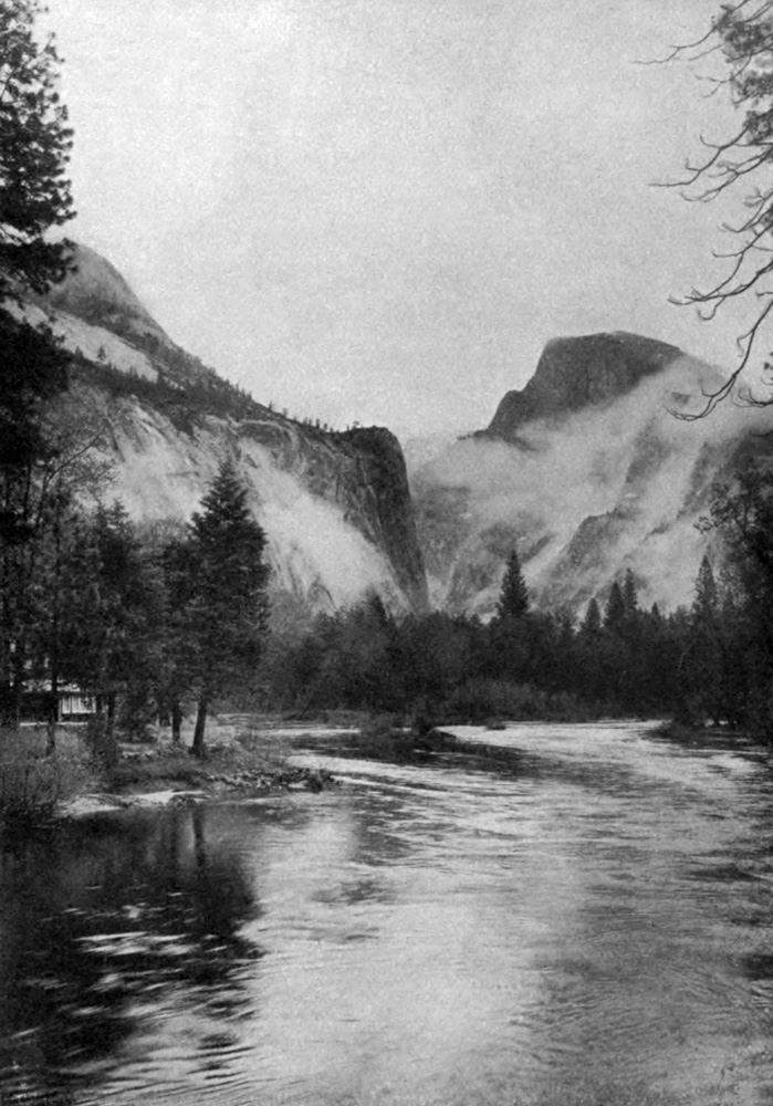 A photograph of a wide, tree-lined river that flows through a valley bordered by massive granite rock domes.