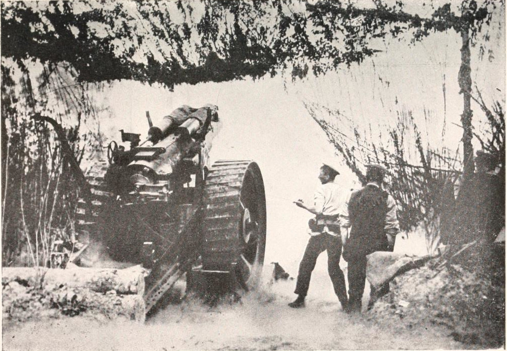 A few men stand next to a large wheeled artillery piece, with a camouflage canopy overhead.