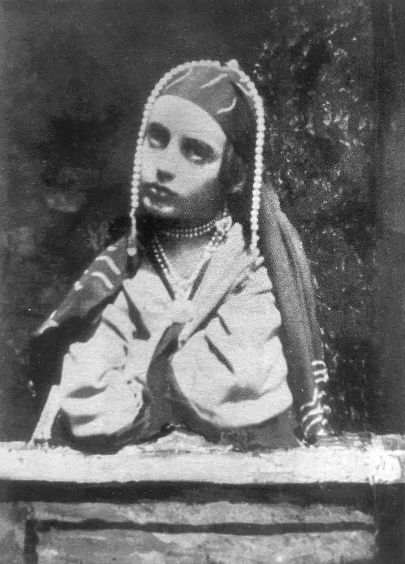 A girl in skullcap with pearls wearing an embroidered shawl and robes leans forward on crossed arms on a cabinet.
